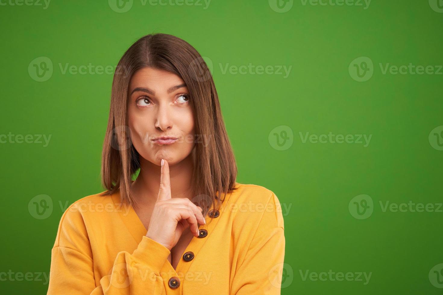 Cerca de mujer en suéter amarillo pensando y apuntando sobre fondo verde foto