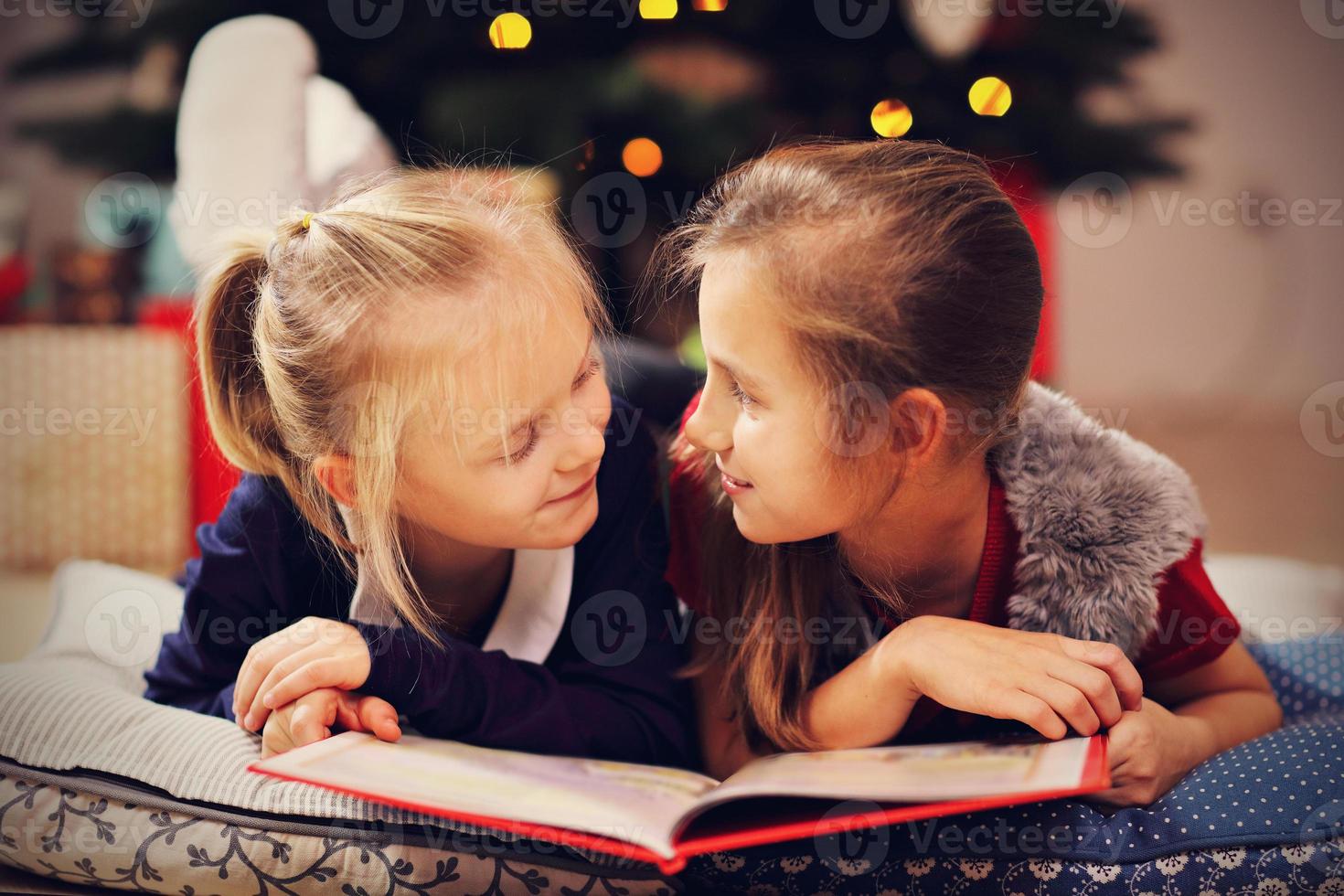 Two cute little sisters reading story book together under Christmas tree photo