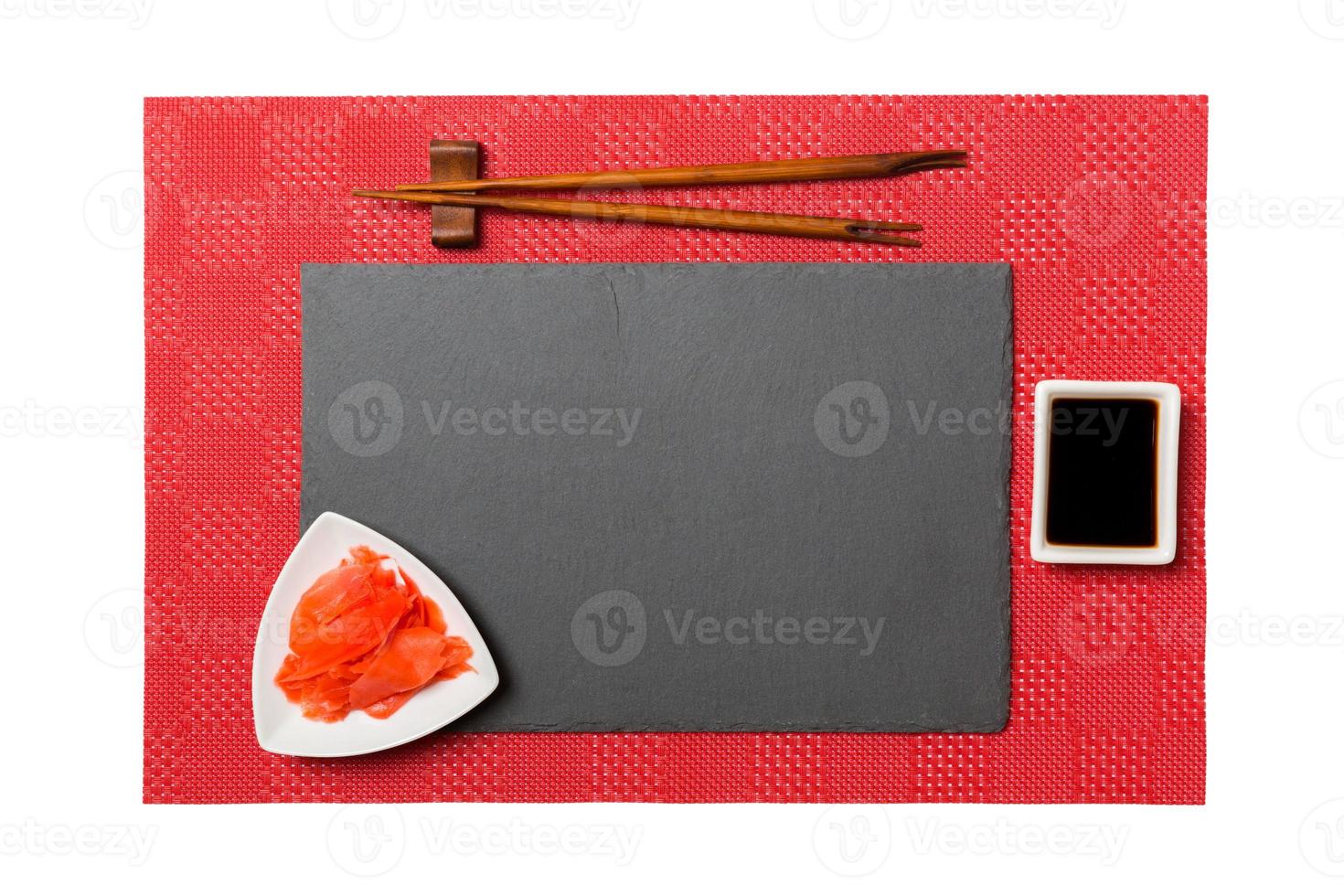 Empty rectangular black slate plate with chopsticks for sushi, ginger and soy sauce on red mat sushi background. Top view with copy space for you design photo