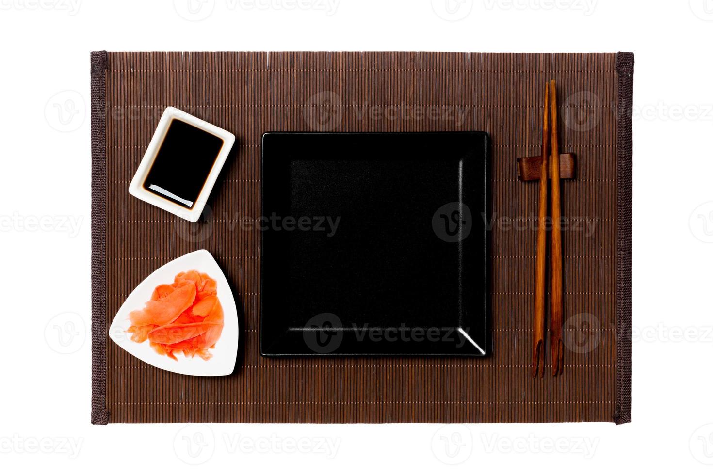 Empty black square plate with chopsticks for sushi, ginger and soy sauce on dark bamboo mat background. Top view with copy space for you design photo
