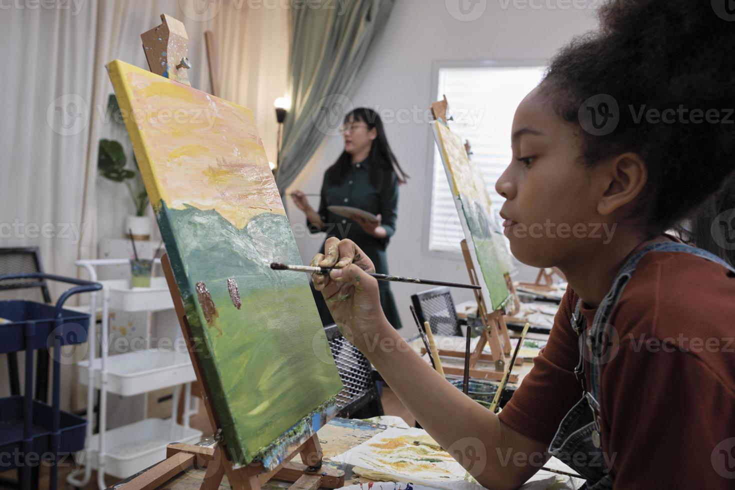 African American girl concentrates on acrylic color picture painting on canvas with students group in art classroom, creative learning with talents and skills in elementary school studio education. photo