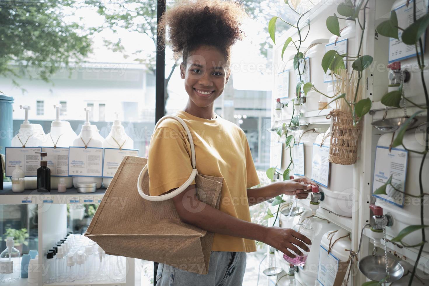 A Black female customer fills liquid soap into recycling jar from reusable containers in a zero-waste and refill store, smiles, and looks at camera, happy shopping in an environment-friendly shop. photo