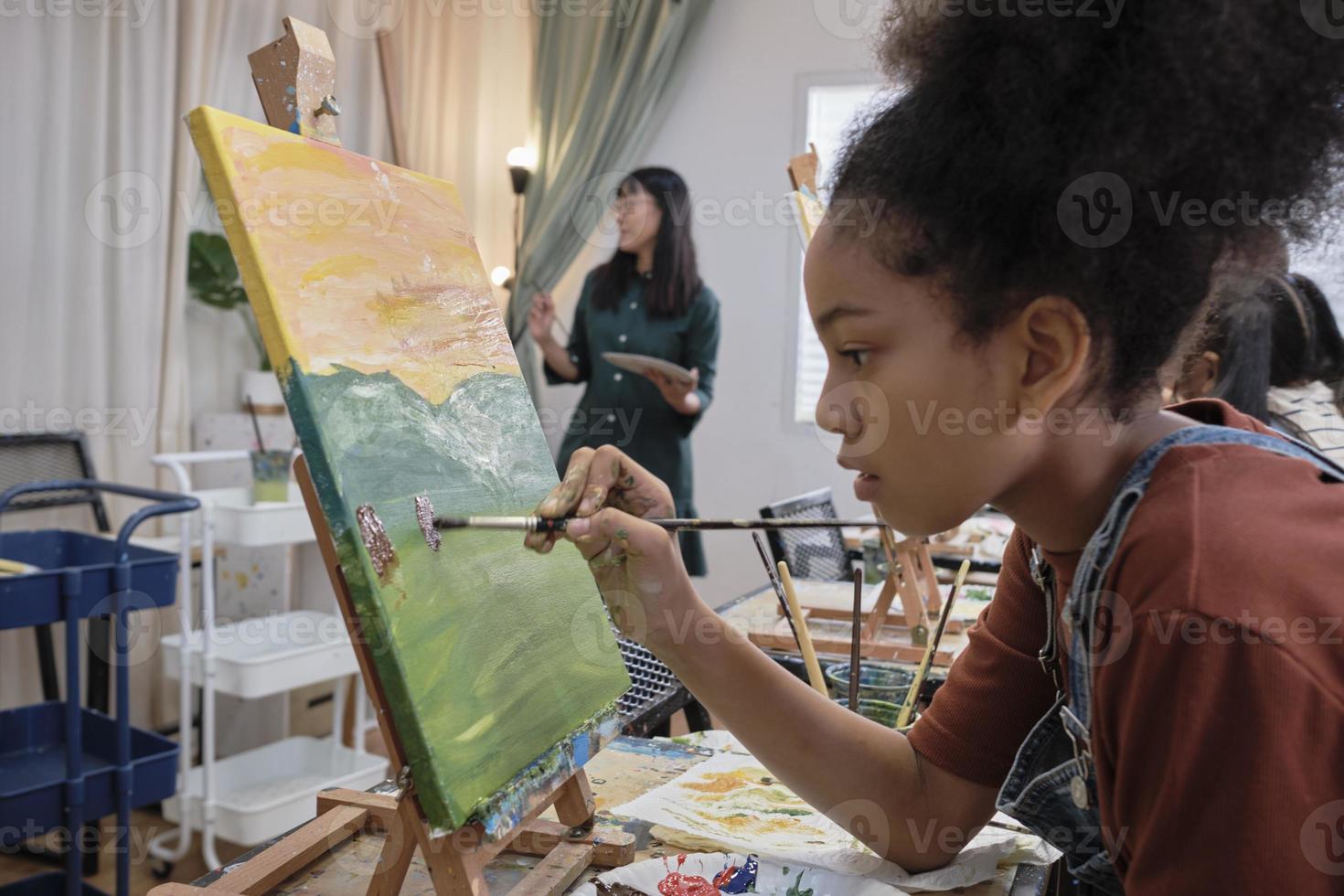 African American girl concentrates on acrylic color picture painting on canvas with students group in art classroom, creative learning with talents and skills in elementary school studio education. photo