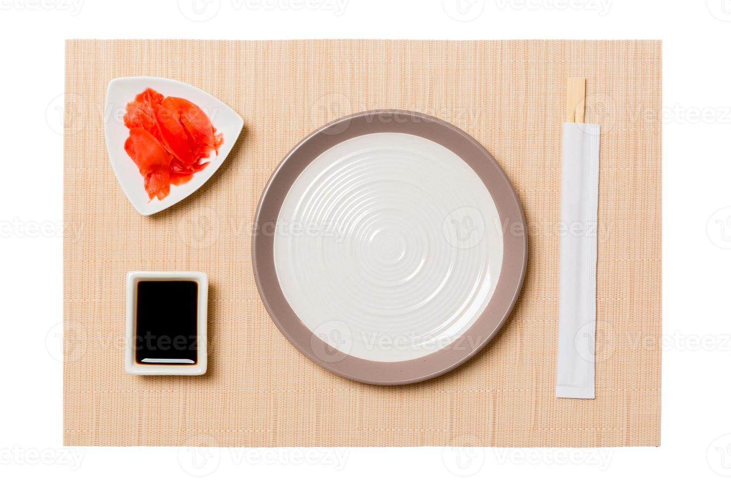 Empty round white plate with chopsticks for sushi, ginger and soy sauce on brown sushi mat background. Top view with copy space for you design photo