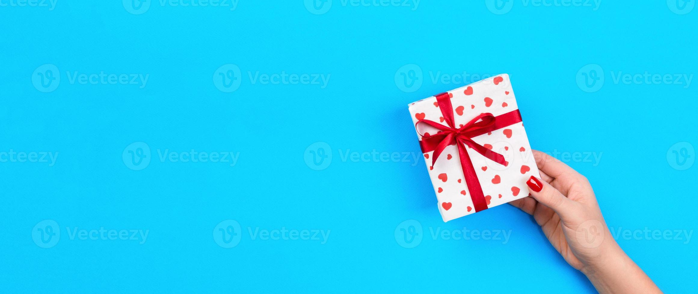 Woman hands give wrapped valentine or other holiday handmade present in paper with red ribbon. Present box, red heart decoration of gift on blue table, Top view banner with copy space for you design photo