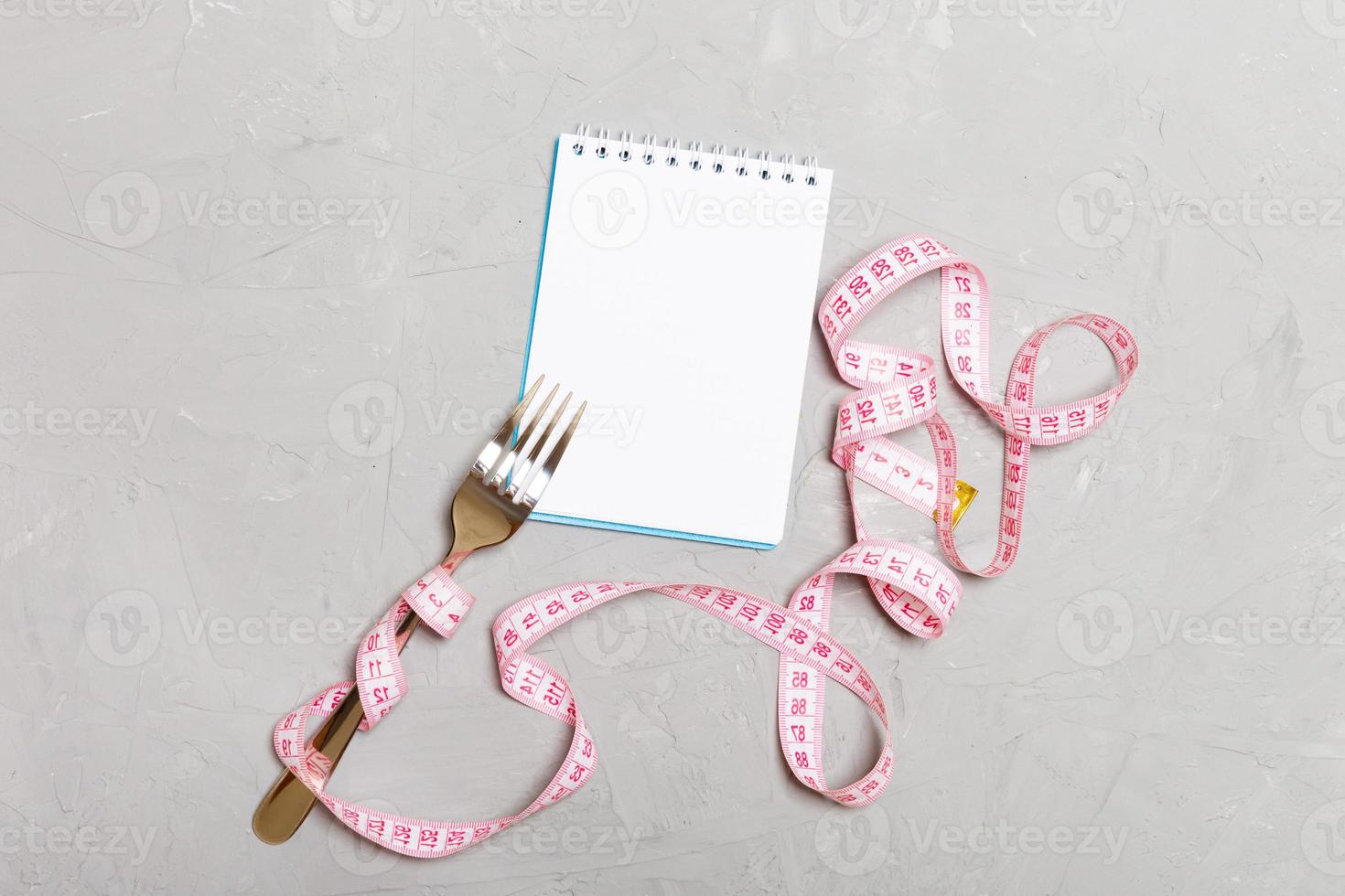 Fitness concept with empty space for your design on cement background. Top view of notepad and fork with pink measure tape photo