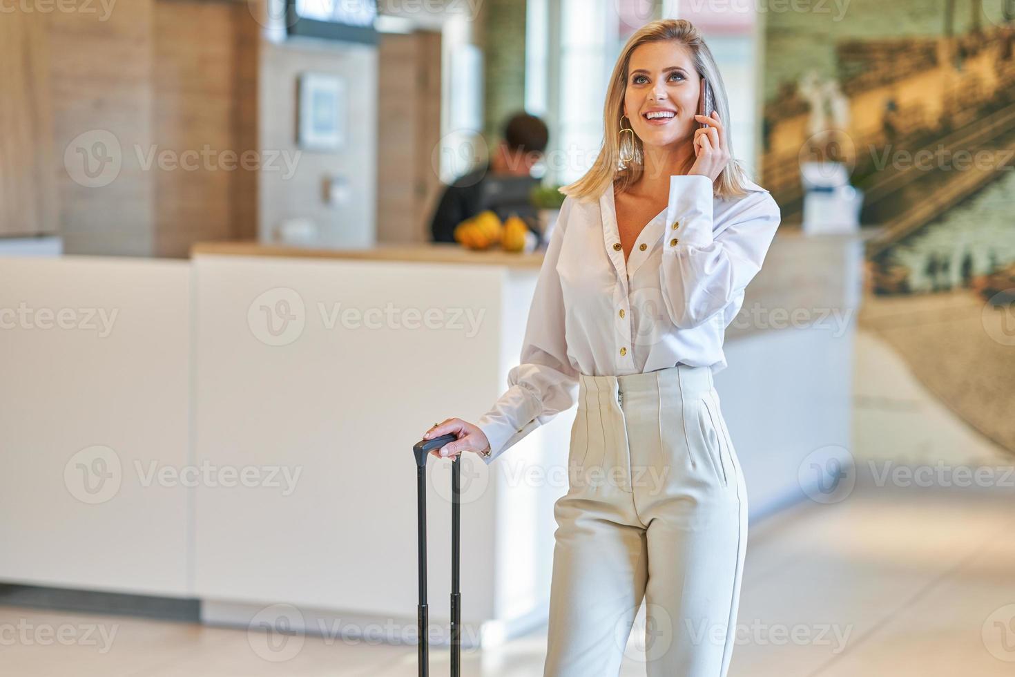 Businesswoman with luggage in modern hotel lobby using smartphone photo