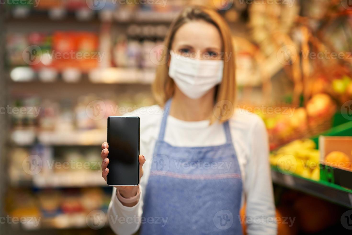 asistente de tienda en máscara médica sosteniendo teléfono inteligente contra tienda de comestibles foto