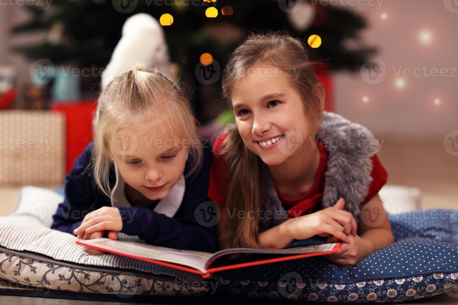 Two cute little sisters reading story book together under Christmas tree photo