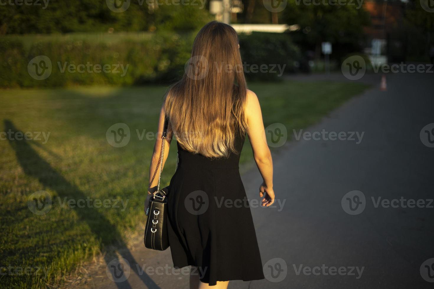 Girl in black dress walks through park. Girl with long hair in summer on street. Black dress and handbag. photo