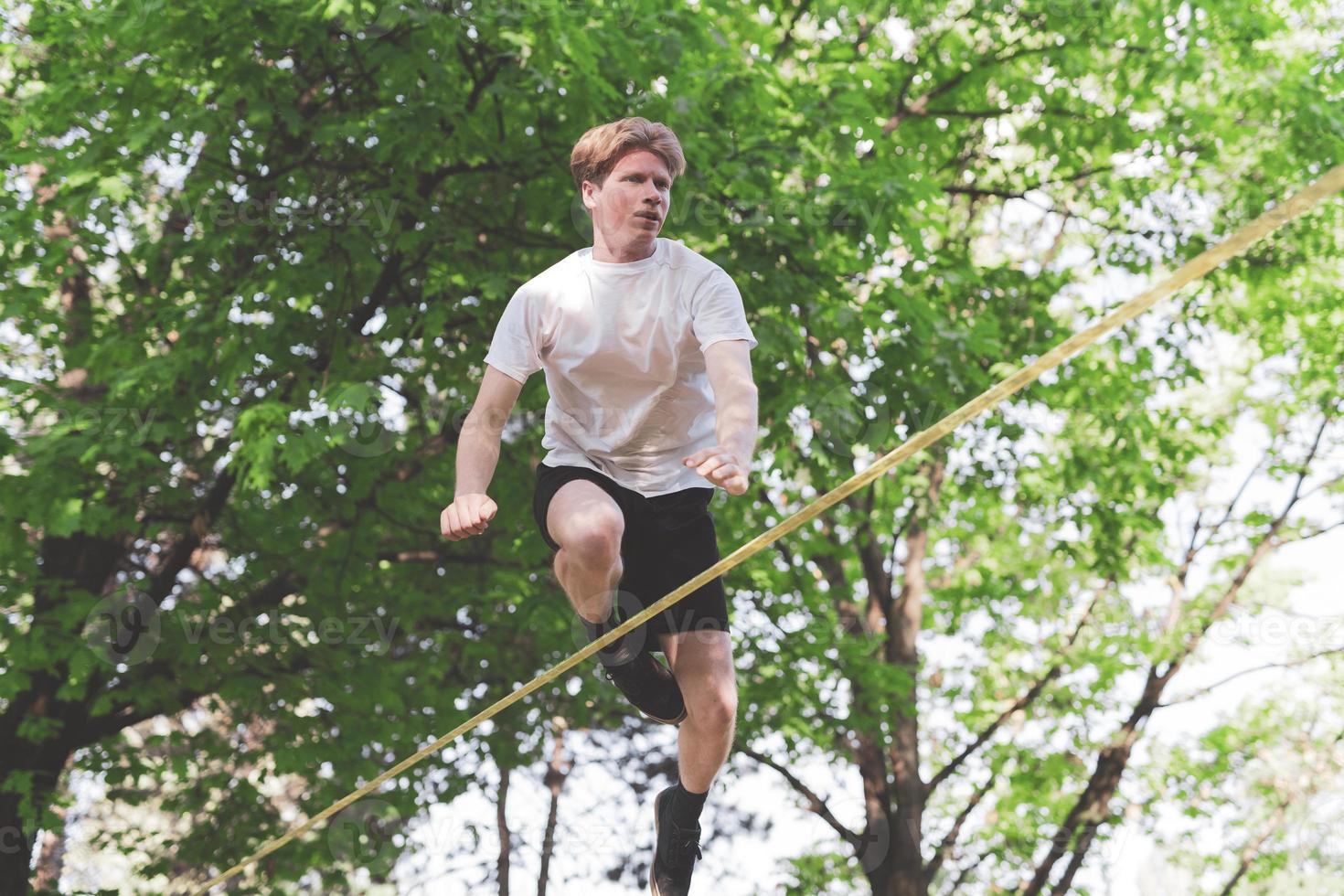 Young man balancing and jumping on slackline. Man walking, jumping and balancing on rope in park. photo