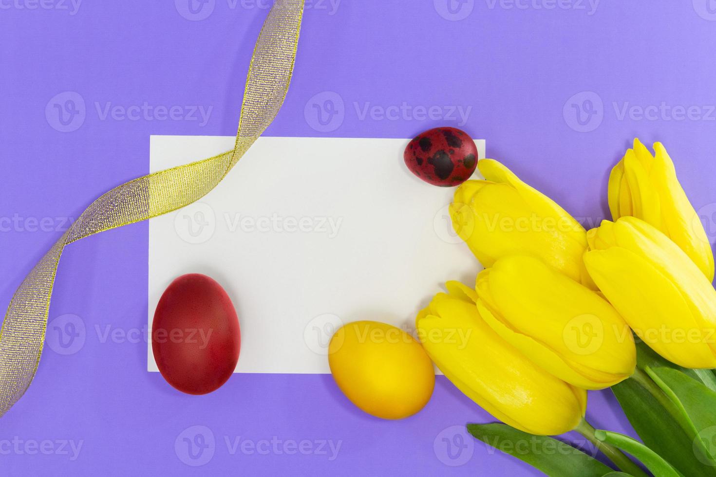 Yellow tulips with empty white sheet for text or mockup and painted yellow and red eggs, gold ribbon. Easter background. Copy space photo