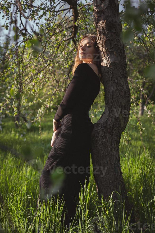 mujer joven en vestido negro apoyado contra el árbol fotografía escénica foto