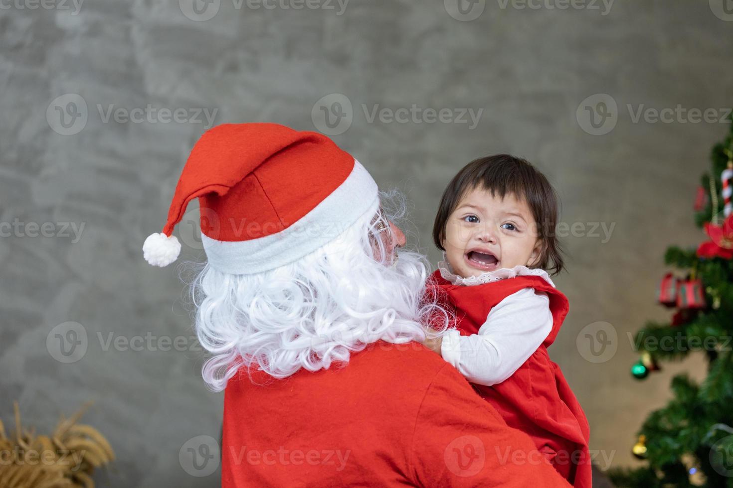 Santa Claus is lifting happy little toddler baby girl up and laughing cheerfully with fully decorated christmas tree on the back for season celebration photo