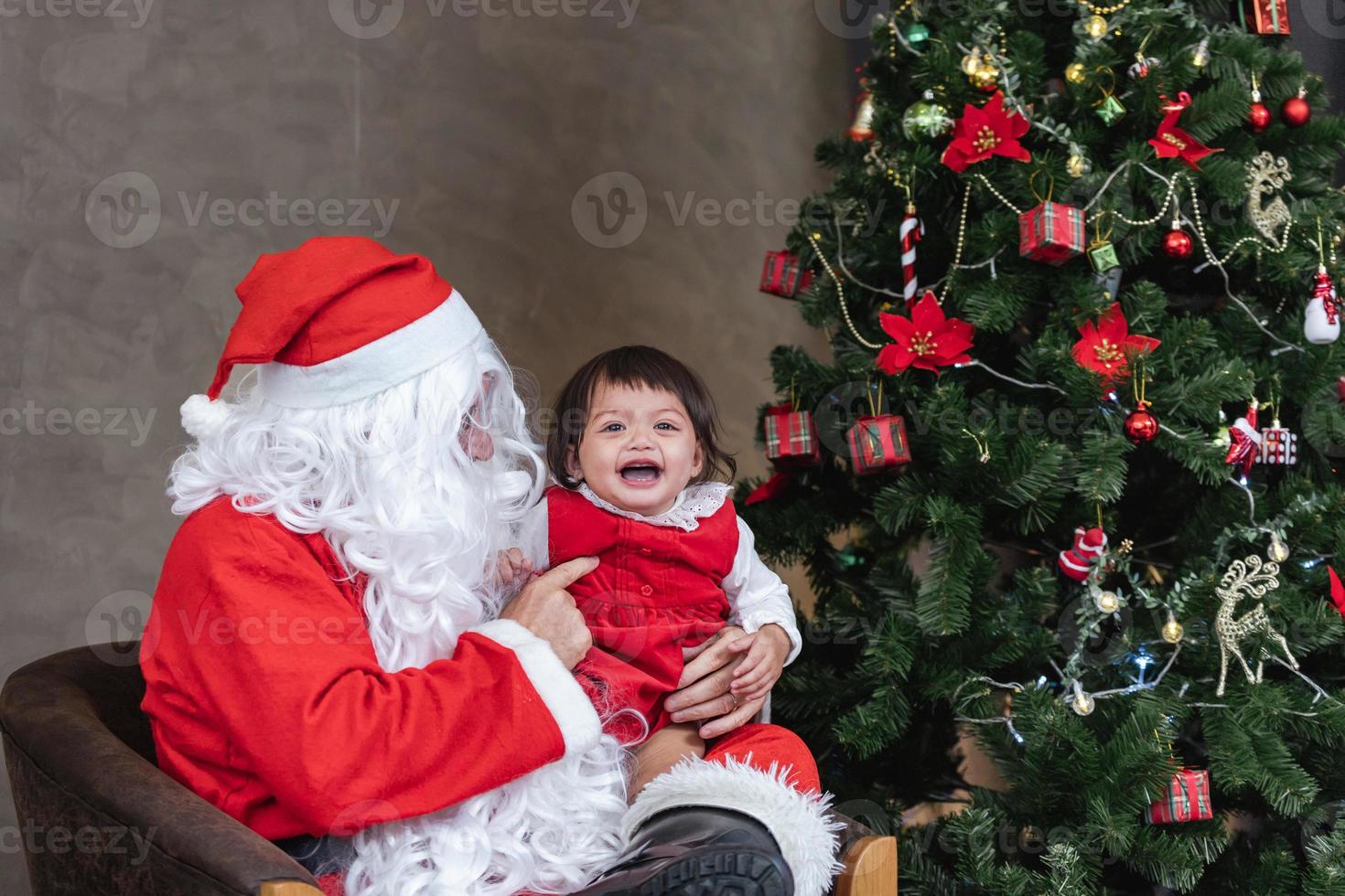 Santa Claus is lifting happy little toddler baby girl up and laughing cheerfully with fully decorated christmas tree on the back for season celebration concept photo