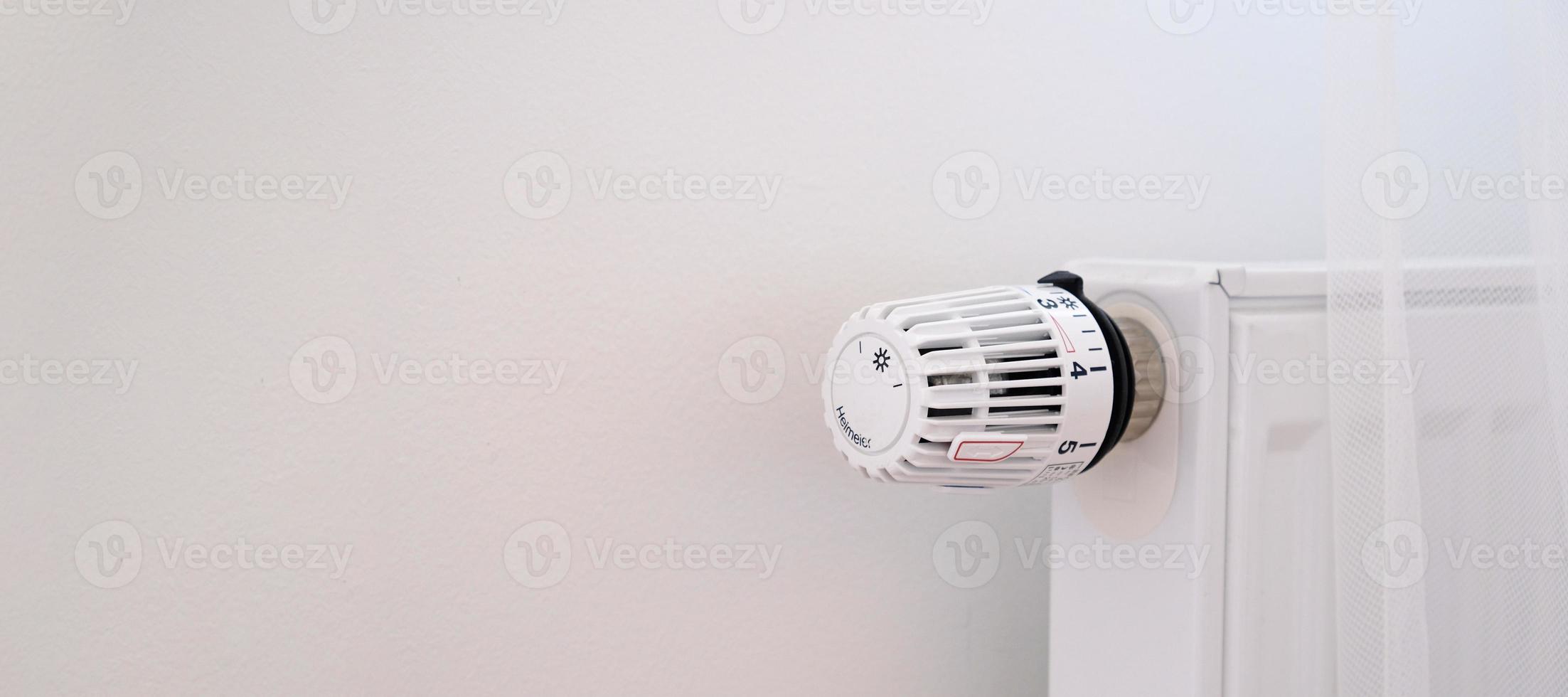 Banner with thermostat and battery temperature controller. Heating service cost concept, utility price increase. Temperature control valve. White heater on wall with curtain in modern apartment. photo