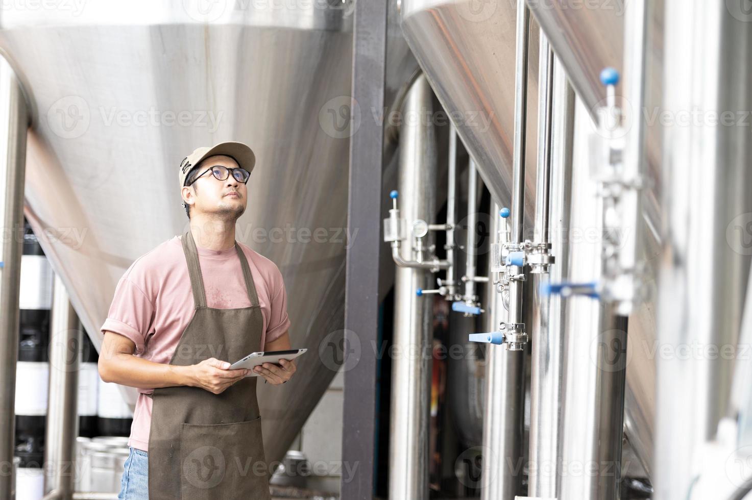 joven trabajador asiático que inspecciona la calidad de la cervecería con un vaso de cerveza artesanal que evalúa la apariencia visual después de la preparación mientras trabaja en una cervecería artesanal de procesamiento. foto