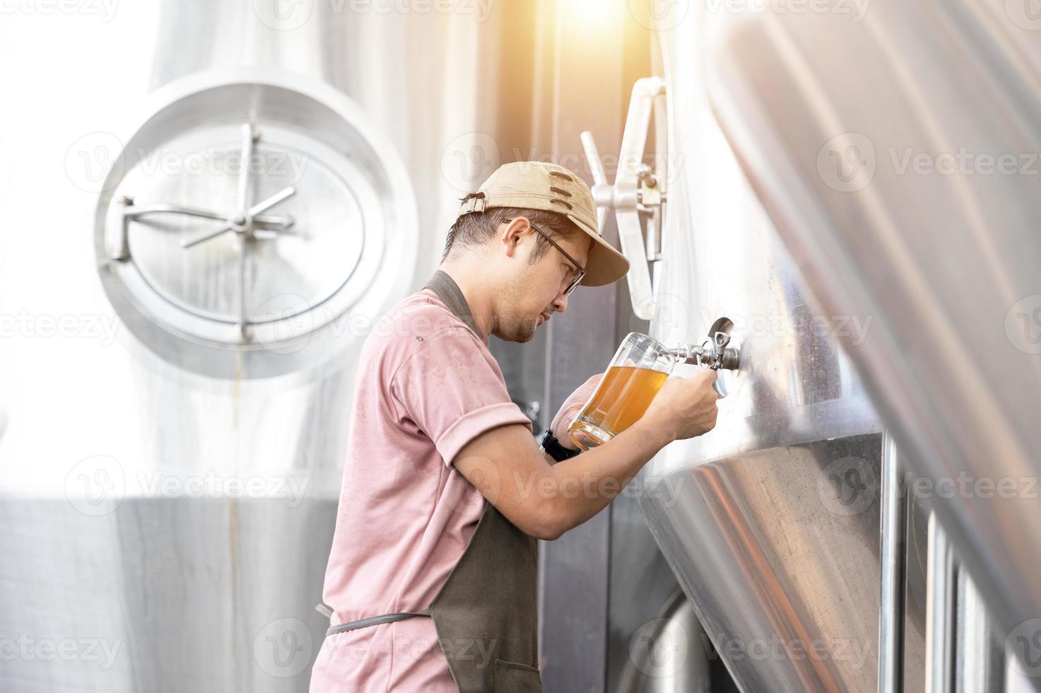 joven trabajador asiático que inspecciona la calidad de la cervecería con un vaso de cerveza artesanal que evalúa la apariencia visual después de la preparación mientras trabaja en una cervecería artesanal de procesamiento. foto