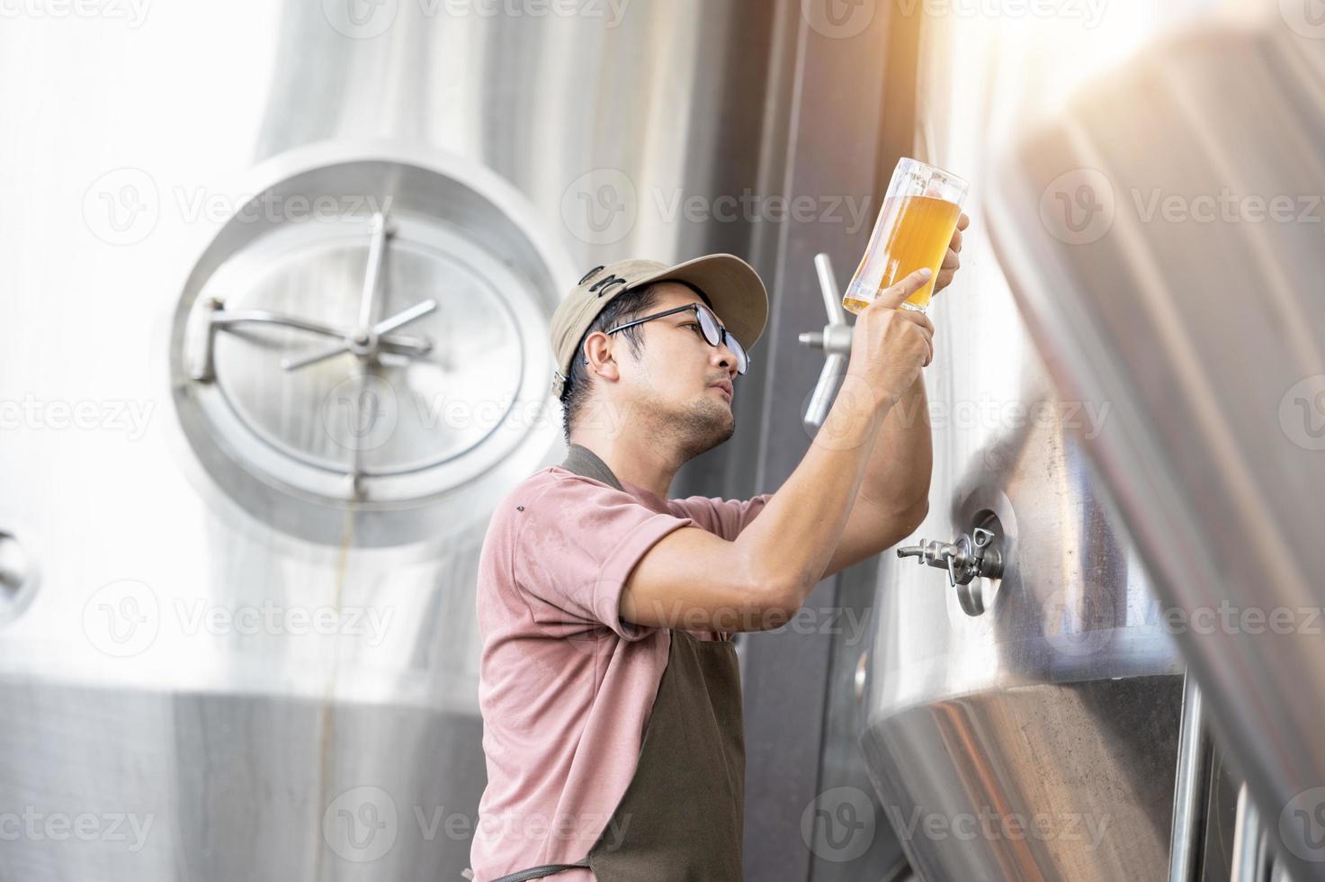 joven trabajador asiático que inspecciona la calidad de la cervecería con un vaso de cerveza artesanal que evalúa la apariencia visual después de la preparación mientras trabaja en una cervecería artesanal de procesamiento. foto