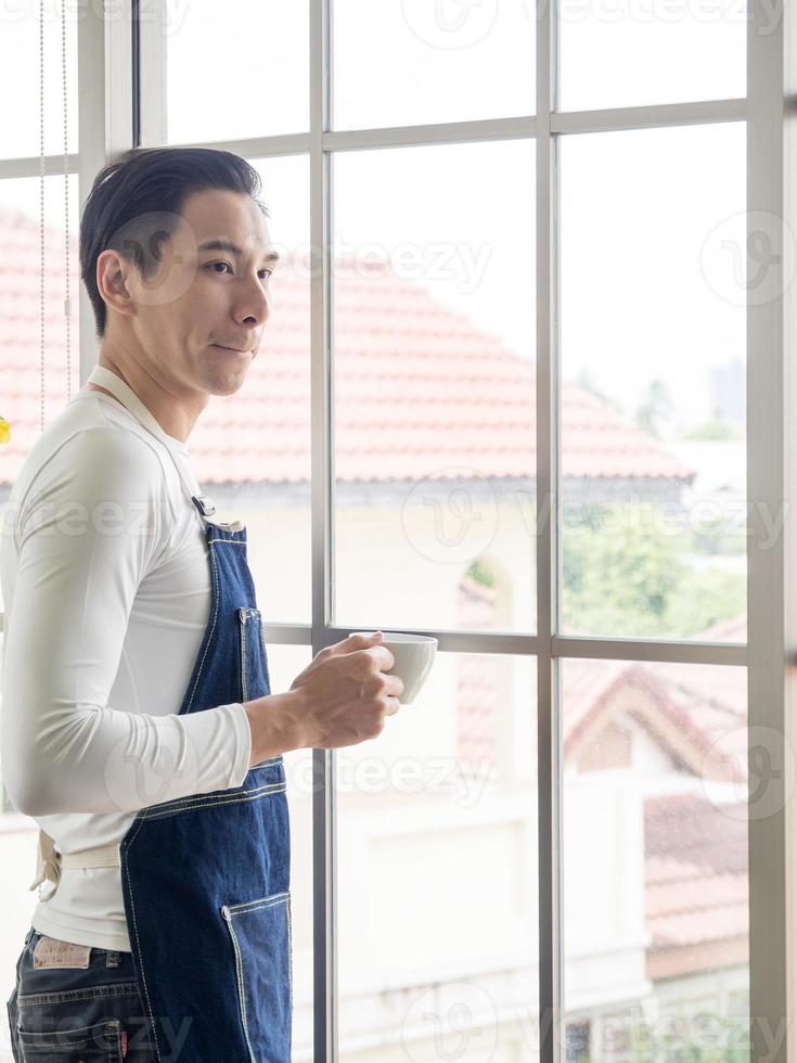 Gardener adult young Asian man one person handsome standing think look and Smiling  relaxing happy drinking a cup of coffee in the room cafe with glass window and green trees in spring morning time. photo