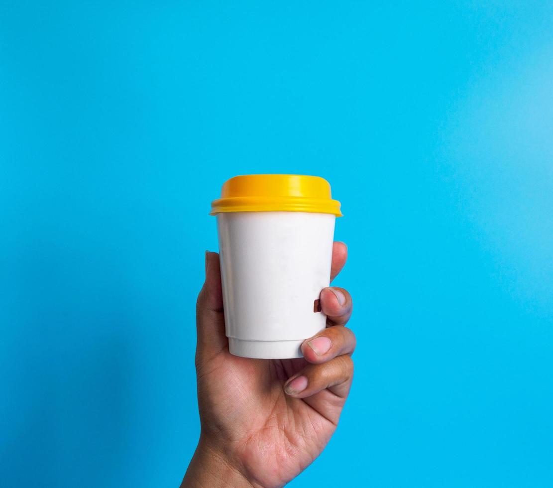 Man hand holding a white peper coffee cup yellow lid, filled with hot black coffee americano no sugar milk ready to drink refreshing. aroma awake fresh to work placed on blue isolated background photo