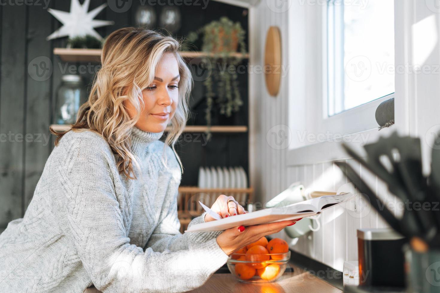 Young woman with blonde curly hair in grey sweater reading book on kitchen at house photo