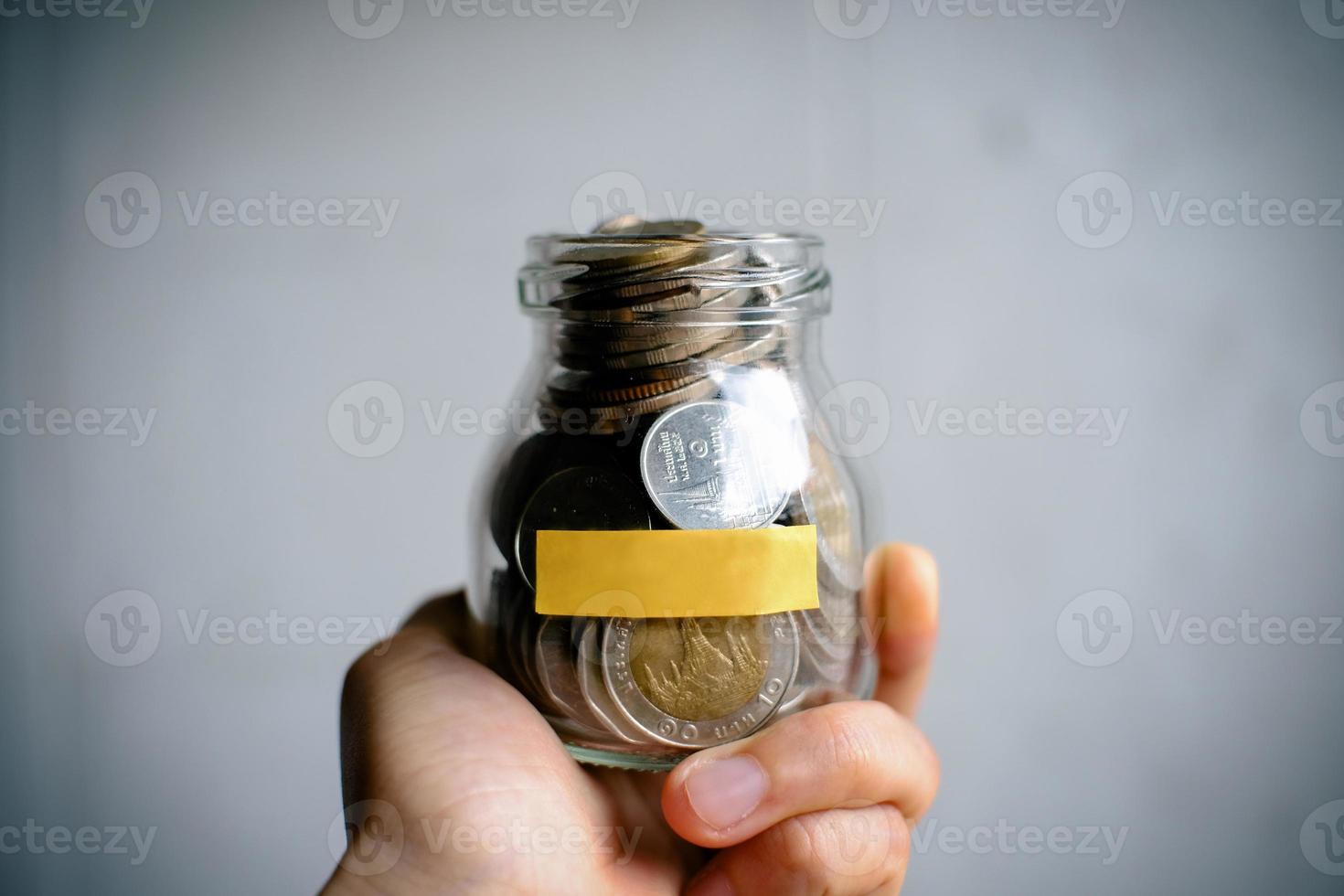 Hand holding money in saving money box with yellow sticky note paper on white background, front view. Stack of Thai Baht coins in a glass jar. photo