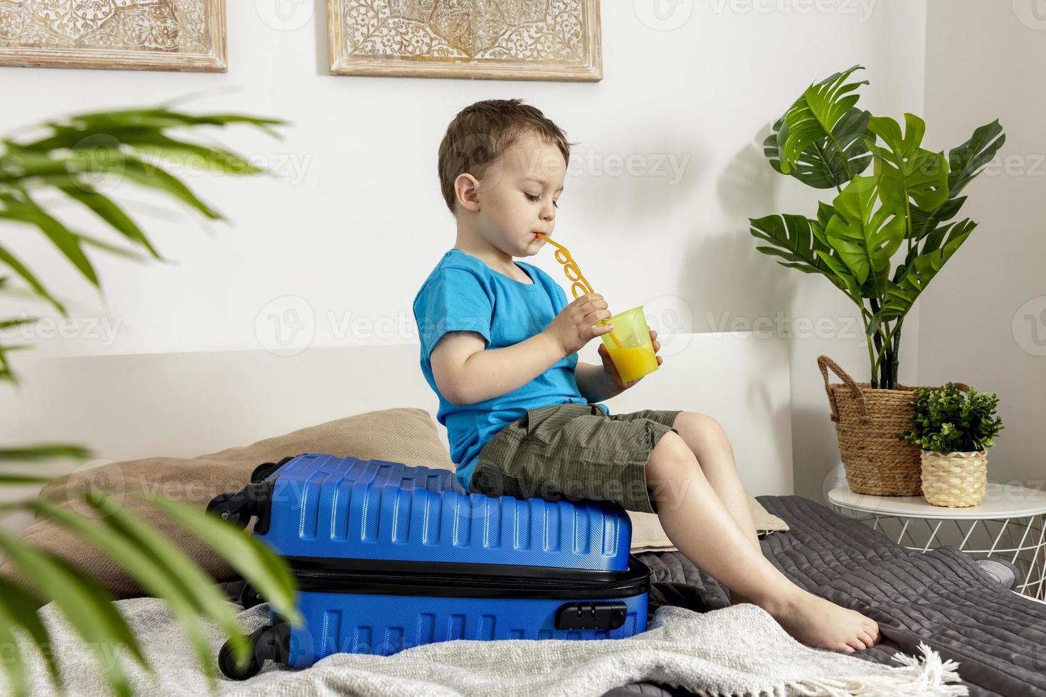 Little caucasian boy with blue shirt ready for vacation. Happy child with blue suitcase going to travel. Tourist, joy of holiday. Kid at home, preparing for flying. Modern and cozy interior. photo