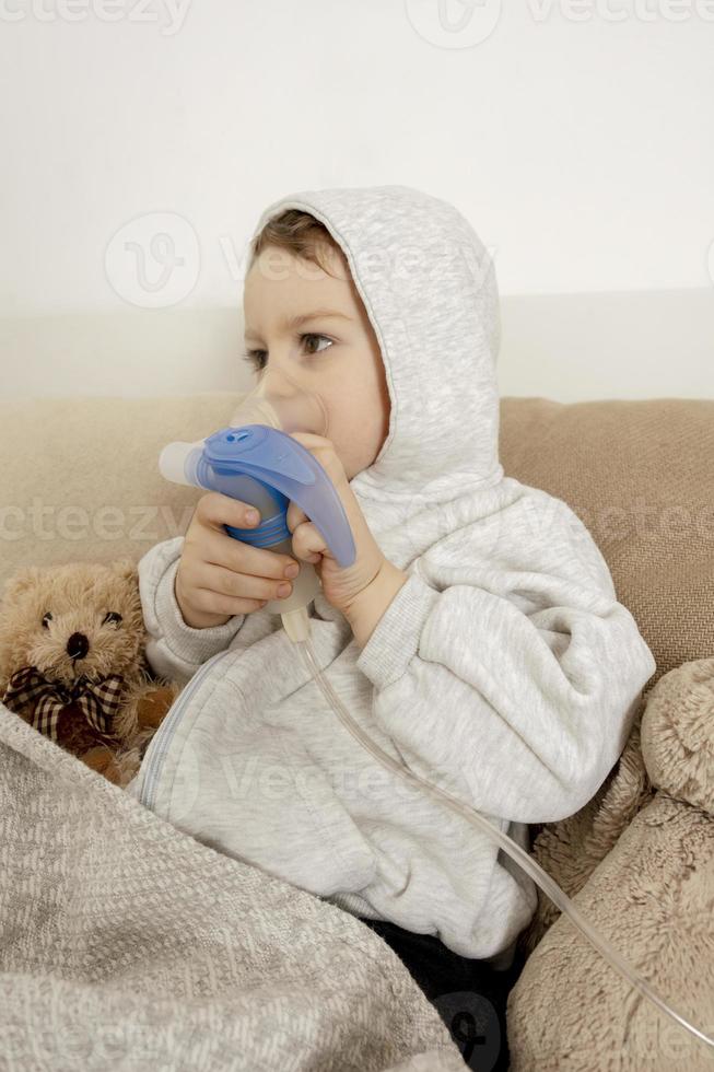 Sick little boy with inhaler for cough treatment. Unwell kid doing inhalation on his bed. Flu season. Medical procedure at home. Interior and clothes in natural earth colors. Cozy environment. photo