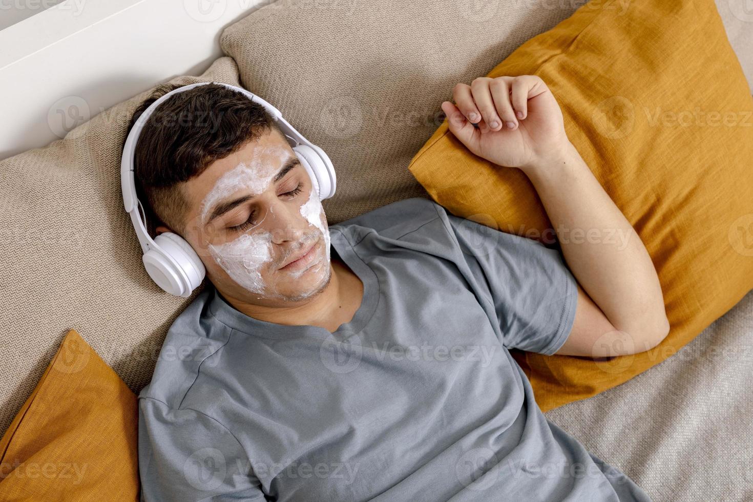 Young man with casual clothes and beauty mask on the face lying on bed at home and resting. Cosmetic for men, skin care. Man listening music or audio book. Relaxing. Time for yourself, mental health. photo