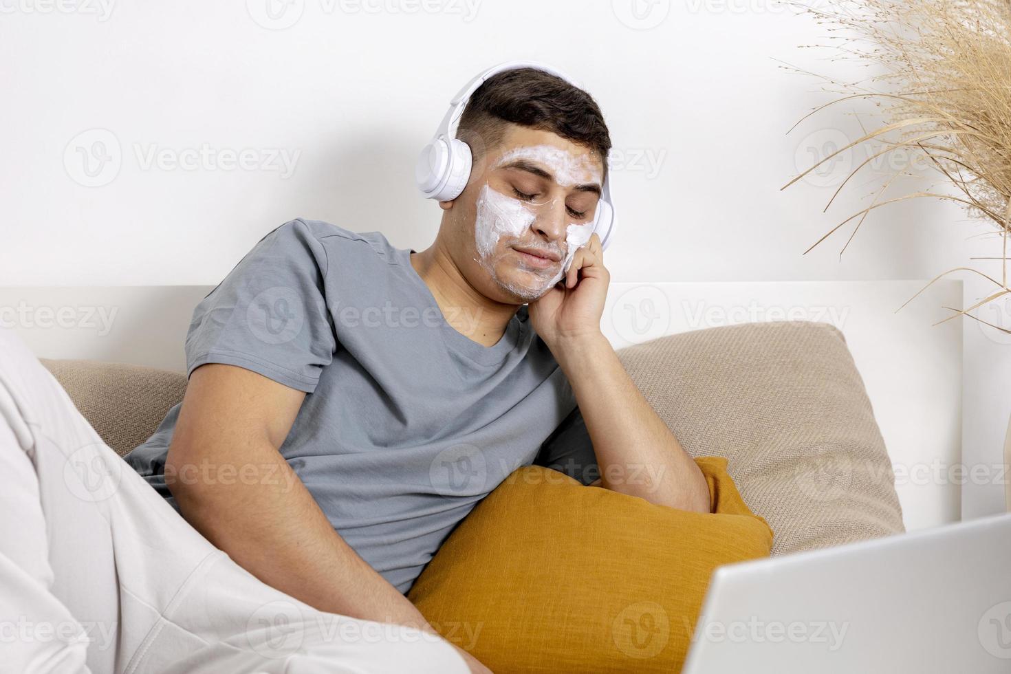 Young man with casual clothes and beauty mask on the face lying on bed with laptop computer and resting. Cosmetic for men, skin care. Man listening music or audio book. Relaxing at home. photo