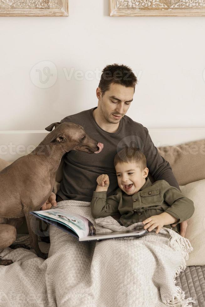 Father and son reading a book on the bed at home. Young attractive man and little boy with dog resting in bedroom. Natural earth colors. Cozy environment. Father reads a fairy tale for his child. photo