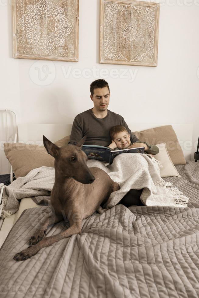Father and son reading a book on the bed at home. Young attractive man and little boy with dog resting in bedroom. Natural earth colors. Cozy environment. Father reads a fairy tale for his child. photo