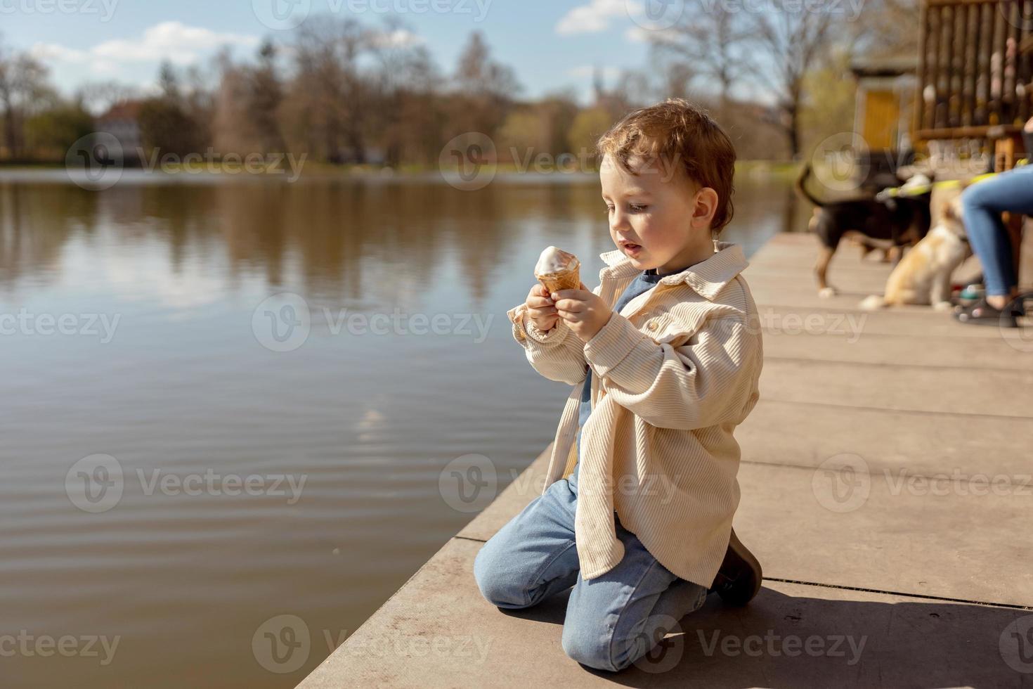 Little adorable boy sitting outdoors and eating ice cream. Lake, water and sunny weather. Child and sweets, sugar. Kid enjoy a delicious dessert. Preschool child with casual clothing. Positive emotion photo