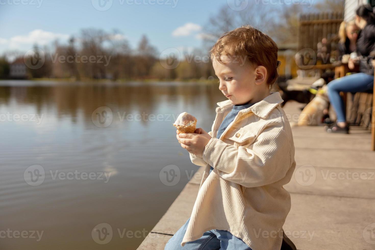 Little adorable boy sitting outdoors and eating ice cream. Lake, water and sunny weather. Child and sweets, sugar. Kid enjoy a delicious dessert. Preschool child with casual clothing. Positive emotion photo