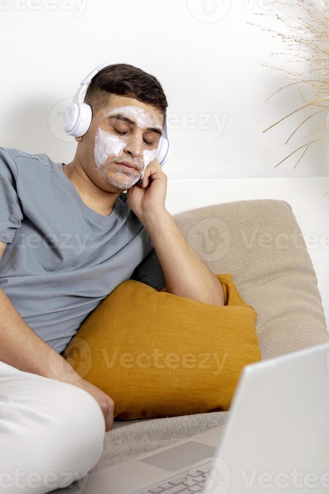 Young man with casual clothes and beauty mask on the face lying on bed with laptop computer and resting. Cosmetic for men, skin care. Man listening music or audio book. Relaxing at home. photo