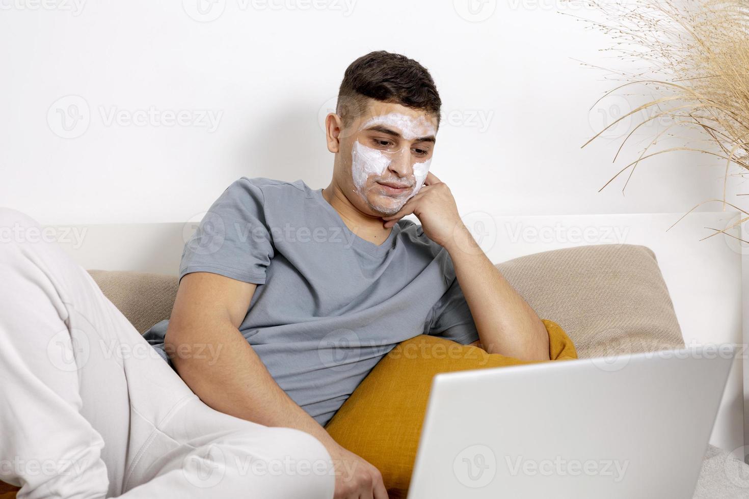 Young man with casual clothes and beauty mask on the face lying on bed with laptop computer and resting. Cosmetic for men, skin care. Man reading news in internet or watching movie. Relaxing at home. photo