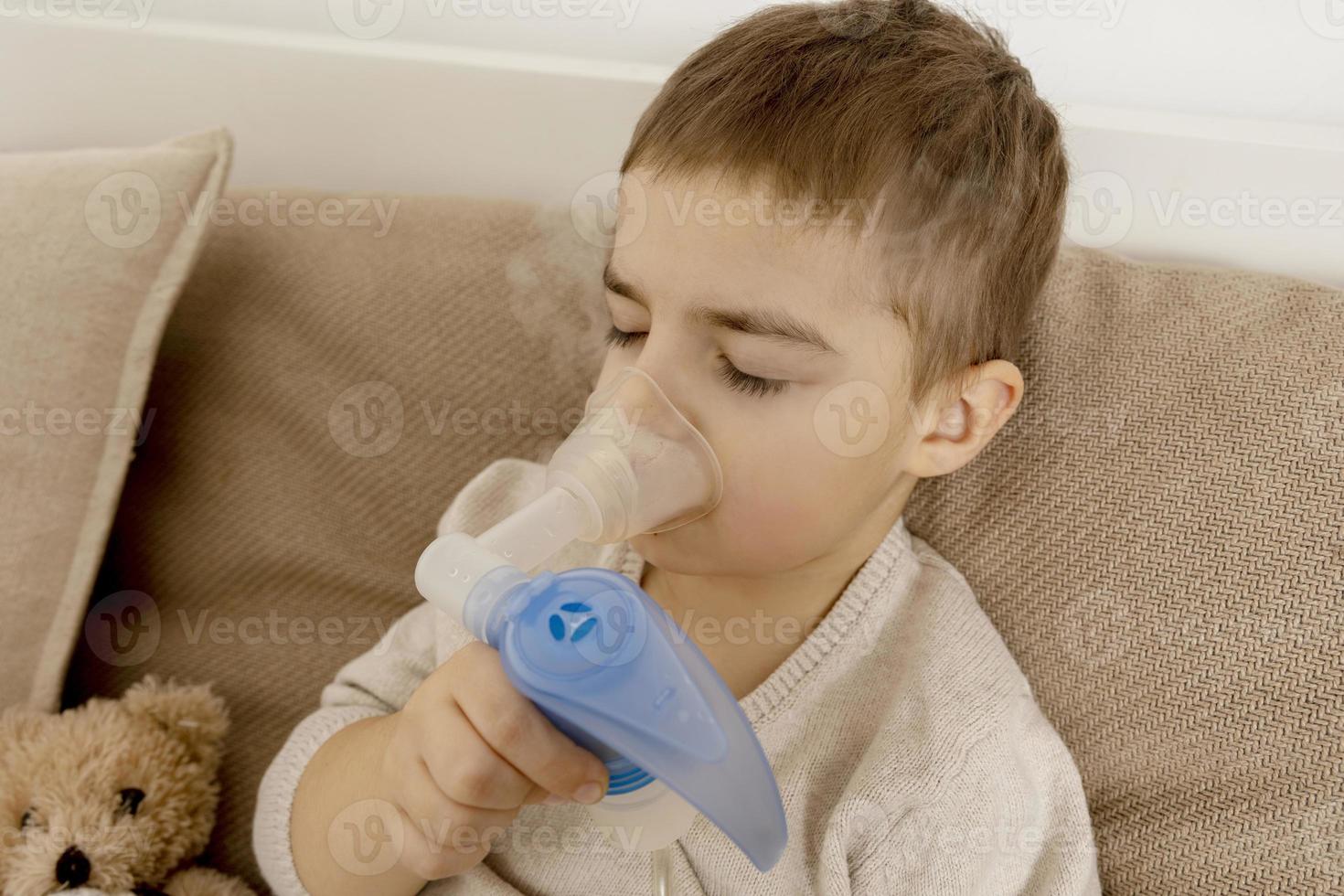 Sick little boy with inhaler for cough treatment. Unwell kid doing inhalation on his bed. Flu season. Medical procedure at home. Interior and clothes in natural earth colors. Cozy environment. photo