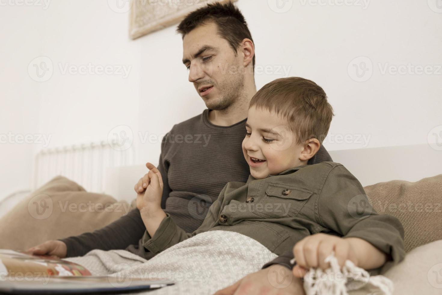 Father and son reading a book on the bed at home. Young attractive man and little boy resting in bedroom. Natural earth colors. Cozy environment. Father reads a fairy tale for his child. photo