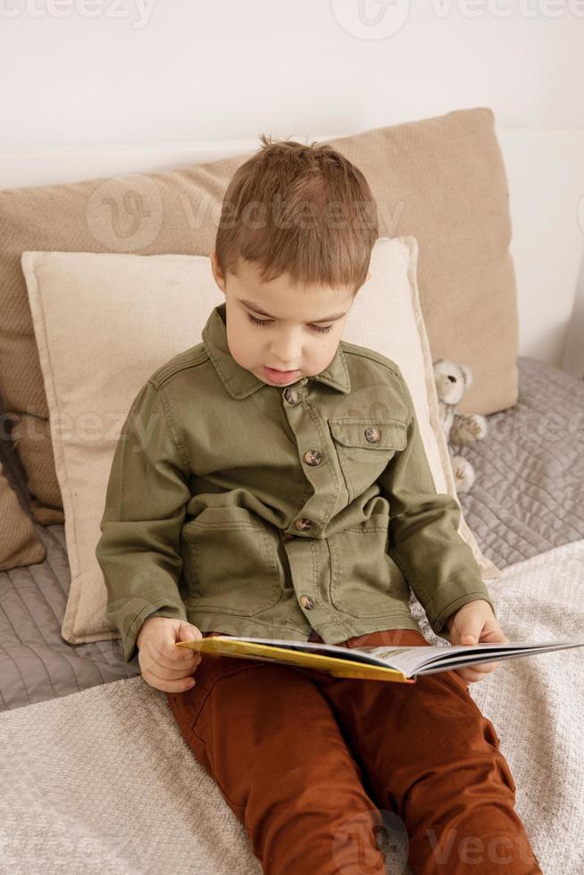 pequeño y lindo niño caucásico leyendo un libro en la cama en casa. interior y ropa en colores tierra naturales. ambiente acogedor. el niño lee un cuento de hadas. foto