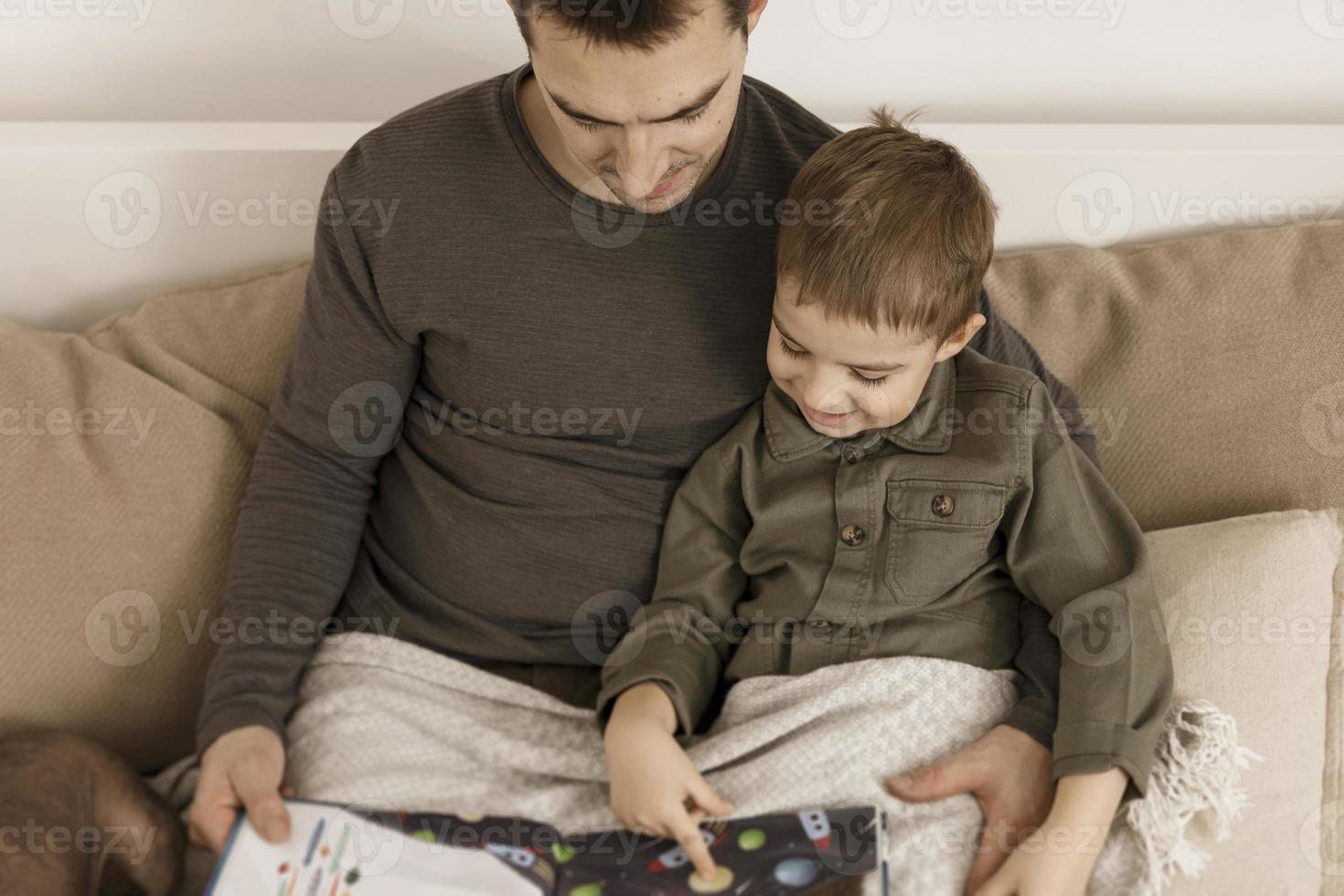 Father and son reading a book on the bed at home. Young attractive man and little boy resting in bedroom. Natural earth colors. Cozy environment. Father reads a fairy tale for his child. photo