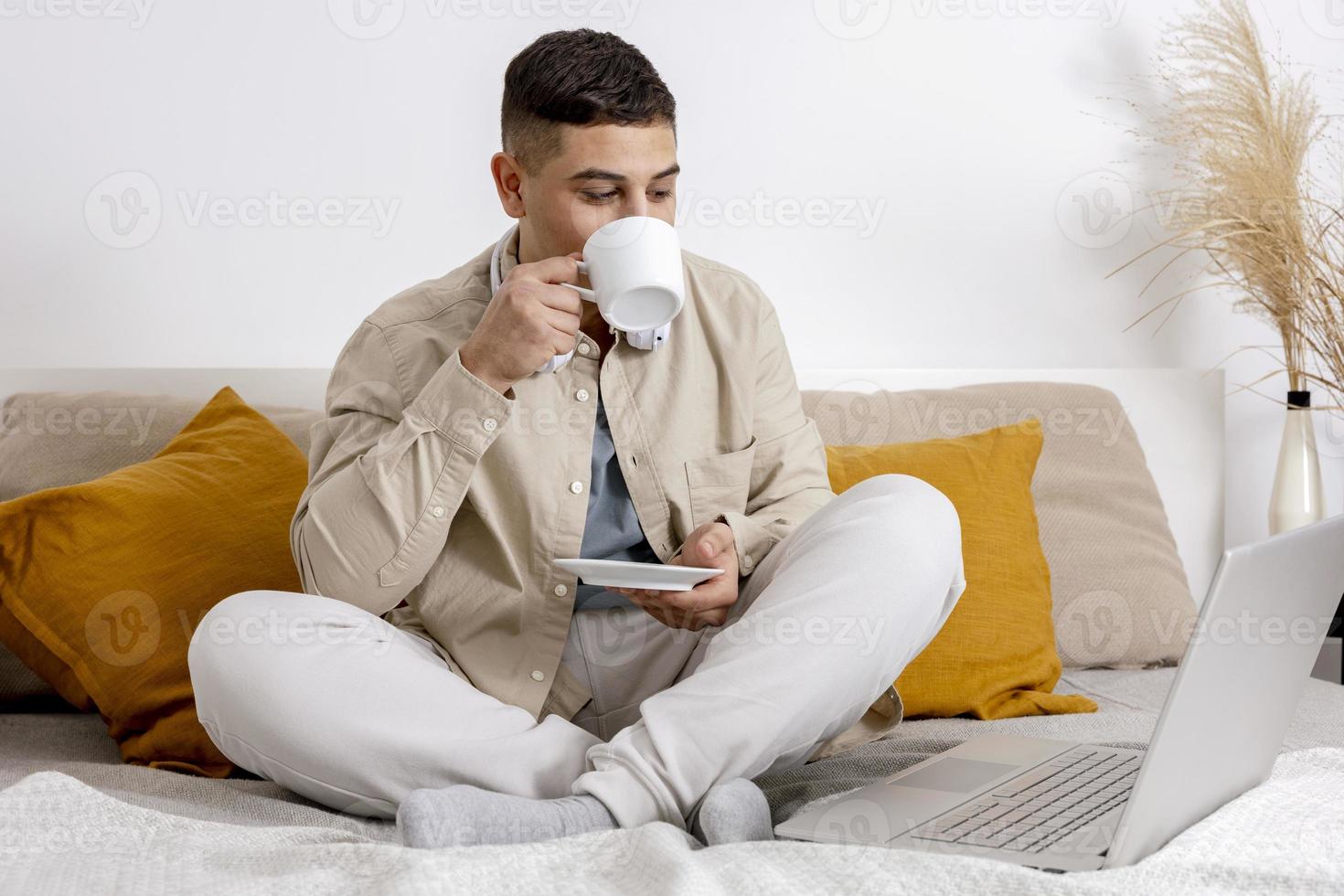 Young man with casual clothes sitting on bed with laptop computer and drinking tea or coffee. Man using notebook to surf in internet, read news, watch movie, call friends. Relaxing at home. photo