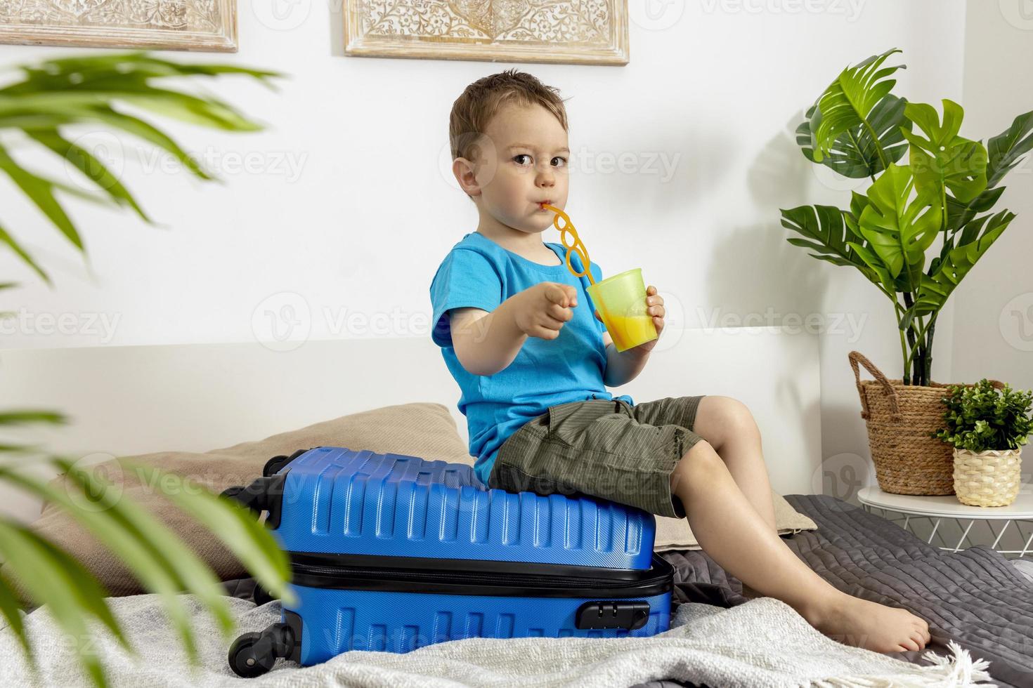 Little caucasian boy with blue shirt ready for vacation. Happy child with blue suitcase going to travel. Tourist, joy of holiday. Kid at home, preparing for flying. Modern and cozy interior. photo
