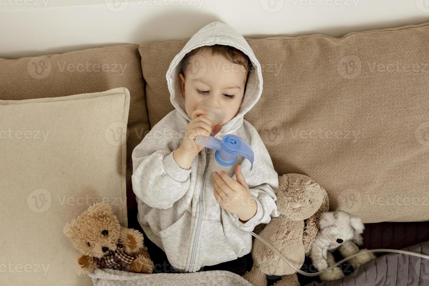 Sick little boy with inhaler for cough treatment. Unwell kid doing inhalation on his bed. Flu season. Medical procedure at home. Interior and clothes in natural earth colors. Cozy environment. photo