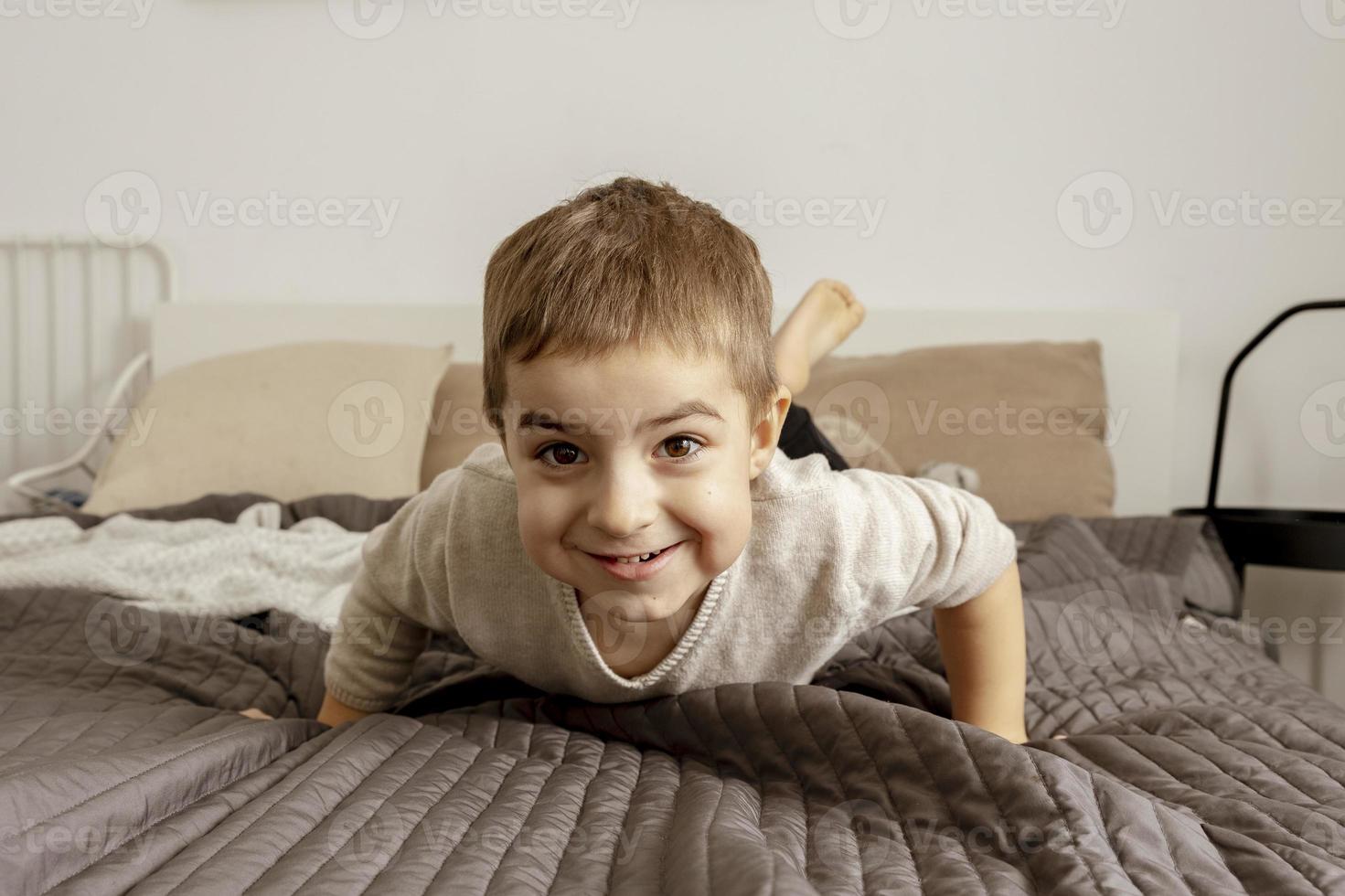 Portrait of little, smiling caucasian boy on the bed at home. Cute child relaxing, resting in bedroom. Positive emotions. Cozy and modern interior. Natural, earth colors. Casual clothes. photo