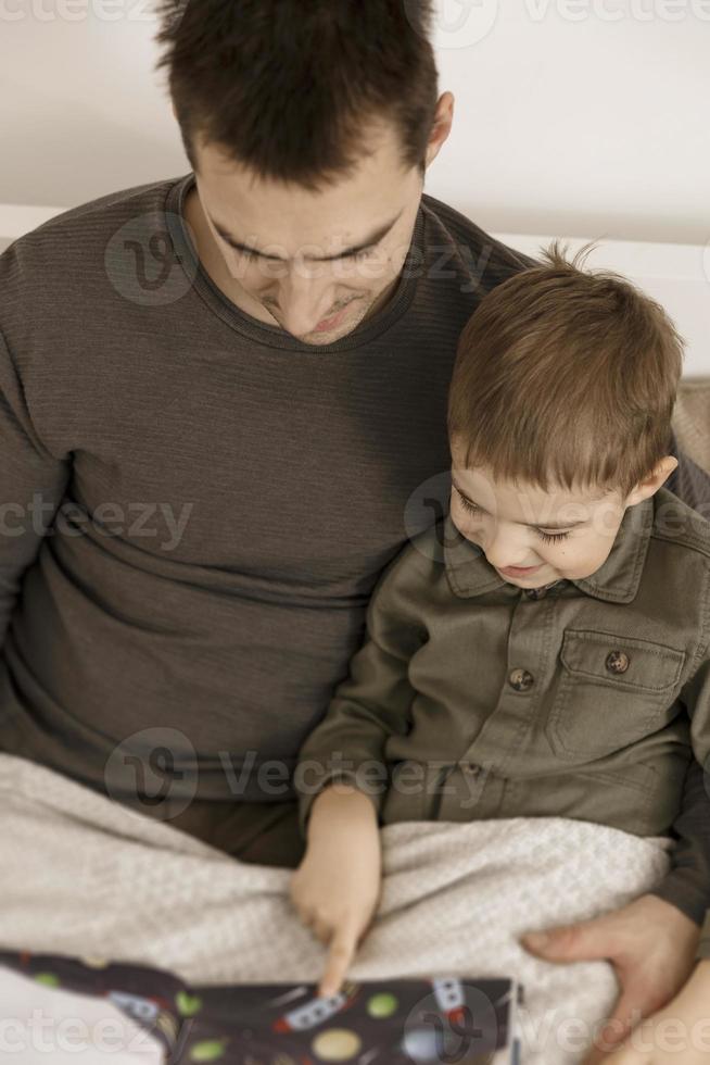 Father and son reading a book on the bed at home. Young attractive man and little boy resting in bedroom. Natural earth colors. Cozy environment. Father reads a fairy tale for his child. photo