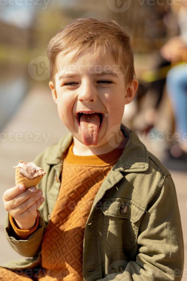 niño adorable sentado al aire libre y comiendo helado. lago, agua y clima soleado. niño y dulces, azúcar. niño disfrute de un delicioso postre. niño en edad preescolar con ropa informal. emoción positiva foto