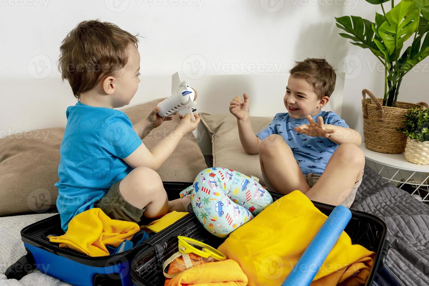 Two little boys with blue shirts ready for vacation. Happy children packing clothes into a suitcase for travel. Tourist, joy of holiday. Kids at home, preparing for flying. Modern and cozy interior. photo