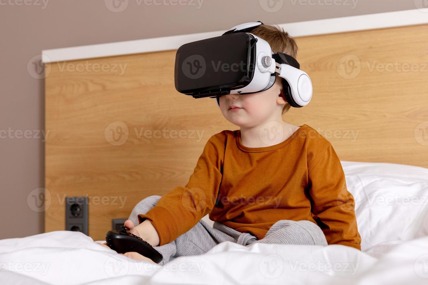 niño adorable sentado en la cama en casa con auriculares vr y jugando  videojuegos interactivos, explorando la realidad virtual. niño lindo con gafas  vr. futuro, gadgets, tecnología, educación en línea. 15940705 Foto