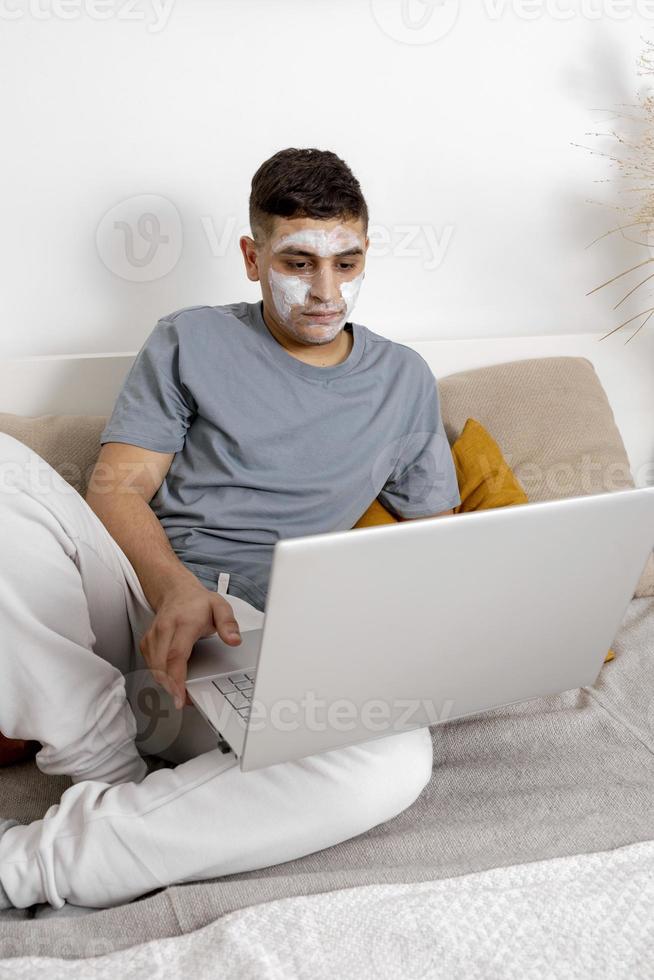 Young man with casual clothes and beauty mask on the face lying on bed with laptop computer and resting. Cosmetic for men, skin care. Man reading news in internet or watching movie. Relaxing at home. photo